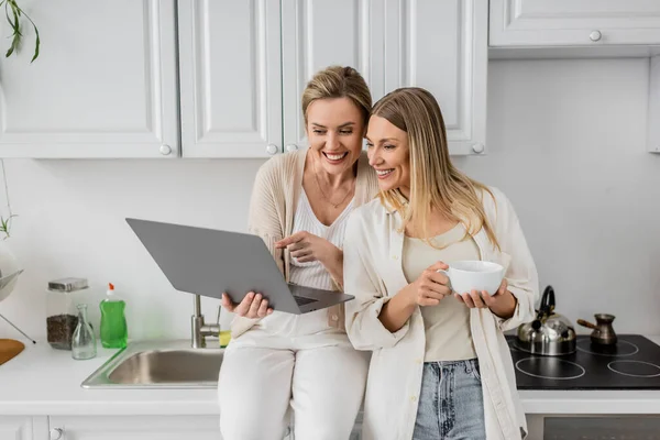 Attractive jolly sisters in lovely outfit looking at laptop on kitchen furniture backdrop, bonding — Stock Photo