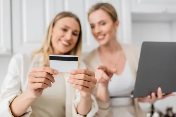 Primer plano bien parecido hermanas de moda en la cocina con el ordenador portátil que sostiene la tarjeta de crédito, borrosa, vinculación — Stock Photo