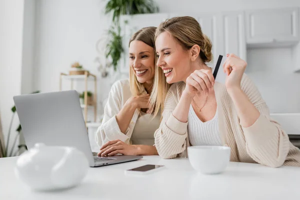 Two cheerful blonde sisters holding credit card looking at laptop and smiling sincerely, bonding — Stock Photo