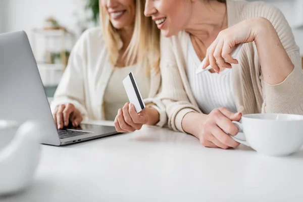 Cropped view of attractive blonde sisters with tea cup and credit card looking at laptop, bonding — Stock Photo