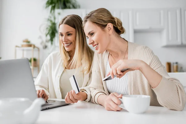 Two lovely blonde sisters in trendy attire with credit card and tea cup looking at laptop, bonding — Stock Photo