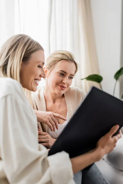 Two smiley attractive sisters in trendy casual cardigans looking at photo book, family bonding — Stock Photo