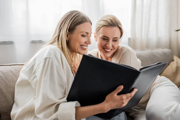 Good looking lovely sisters in casual attire sitting on sofa looking at photo album, family bonding — Stock Photo