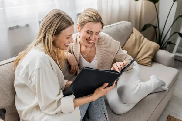 Two beautiful blonde sisters sitting on sofa and looking at photo album, quality time, bonding — Stock Photo