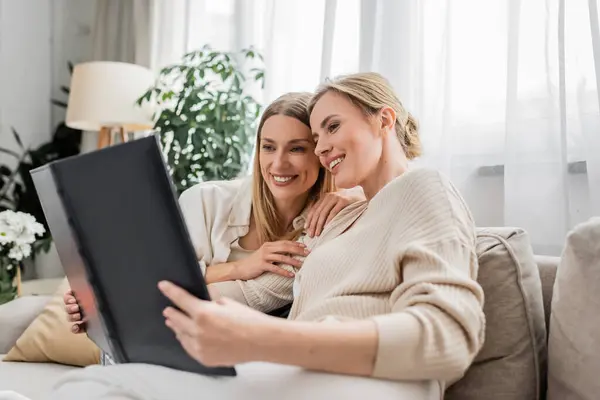 Pretty young sisters hugging and looking at photo book while sitting on sofa, family bonding — Stock Photo