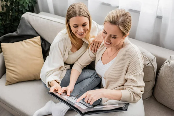 Two cheerful pretty sisters sitting comfortably on sofa looking at photo album, family bonding — Stock Photo