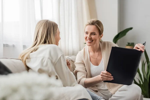 Two cheerful blonde sisters in trended attire looking at each other holding photo album, bonding — Stock Photo