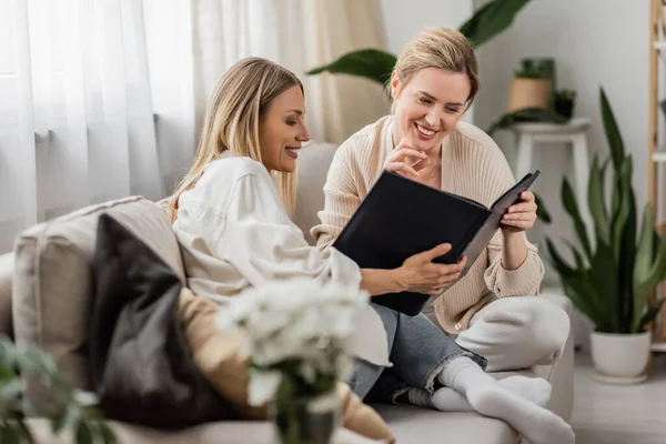 Cheerfully smiling sisters holding photo book in their hands, hand to chin, family bonding — Stock Photo