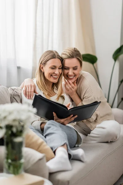 Pretty and lovely sisters in trendy attire laughing and looking at photo album, family bonding — Stock Photo