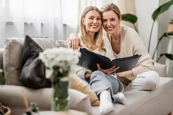 Dos hermanas alegremente atractivas sentadas en el sofá con el álbum de fotos abrazando y mirando a la cámara, vinculación - foto de stock