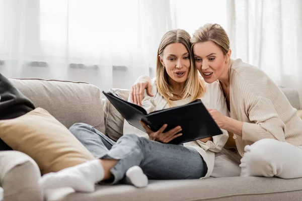 Surprised blonde sisters in trendy outfit looking at photo album on white curtain backdrop, bonding — Stock Photo