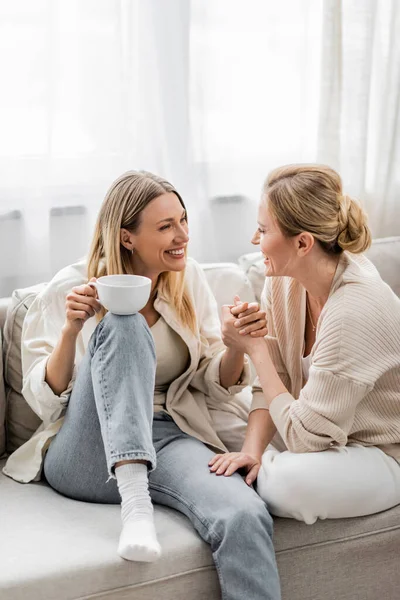 Zwei reizende Schwestern in trendiger Pastellkleidung, die auf dem Sofa sitzen und Tee trinken, Händchen haltend, — Stockfoto
