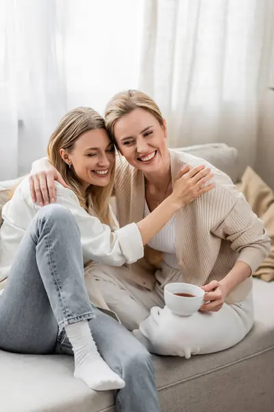Zwei gut aussehende hübsche Schwestern in trendiger Kleidung, die auf dem Sofa sitzen und sich umarmen, Familienbande — Stockfoto