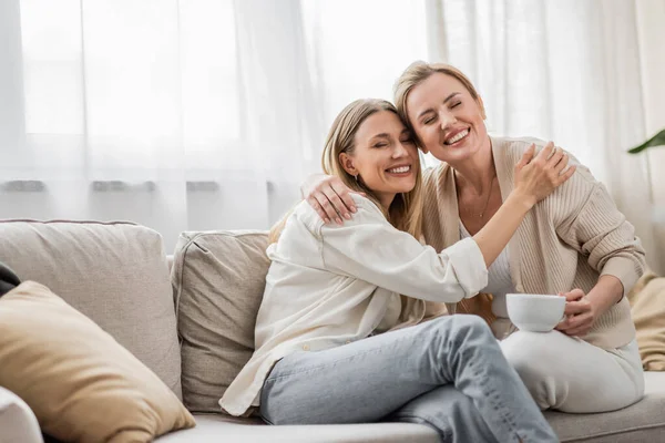 Dos hermanas rubias guapas sentadas en el sofá y abrazadas con los ojos cerrados, unión familiar - foto de stock