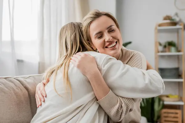 Two blonde sisters in lovely casual attire hugging on sofa with closed eyes, family bonding — Stock Photo