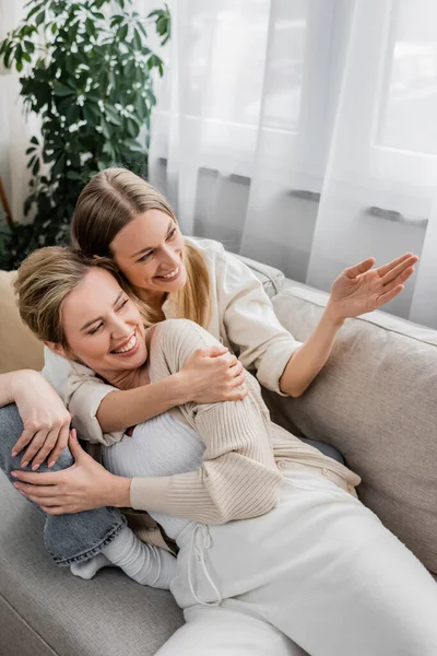 Joyeux soeurs blondes étreignant chaudement sur canapé regardant par la fenêtre, ensemble, liens familiaux — Photo de stock