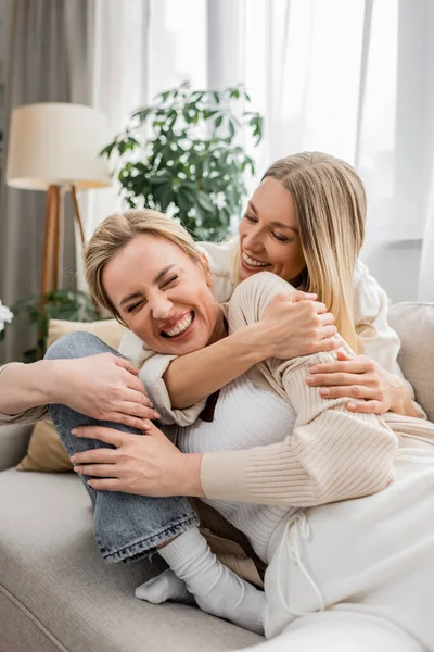 Duas irmãs encantadoras atraentes no sofá rindo e abraçando calorosamente, união, ligação familiar — Fotografia de Stock