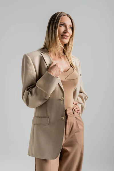 Attraente donna bionda in abito beige e marrone sorridente guardando lontano su sfondo grigio, moda — Foto stock