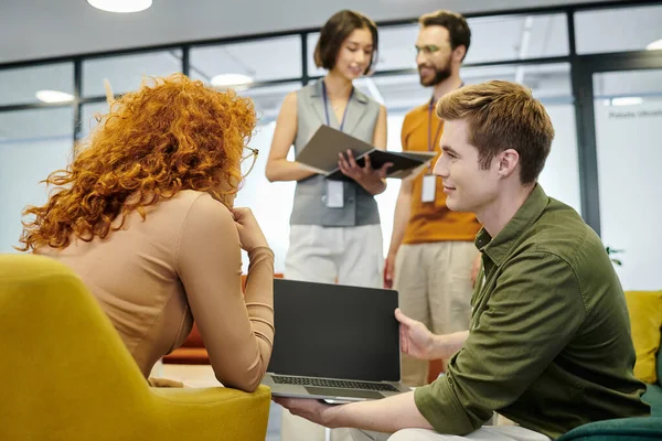 Unternehmer hält Laptop in der Nähe einer rothaarigen Geschäftsfrau, während Kollegen mit Dokumenten arbeiten — Stockfoto