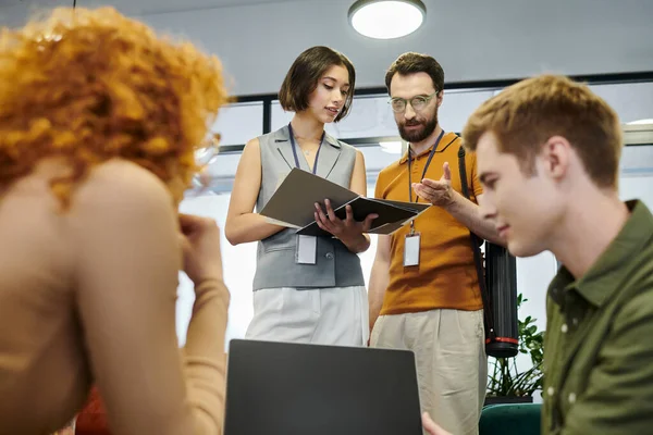 Homme d'affaires barbu pointant vers des documents près de la femme et des collègues flous avec ordinateur portable au bureau — Photo de stock