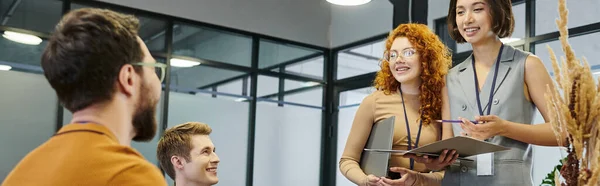 Positive businesswomen with papers and laptop talking to colleagues in office, banner — Stock Photo