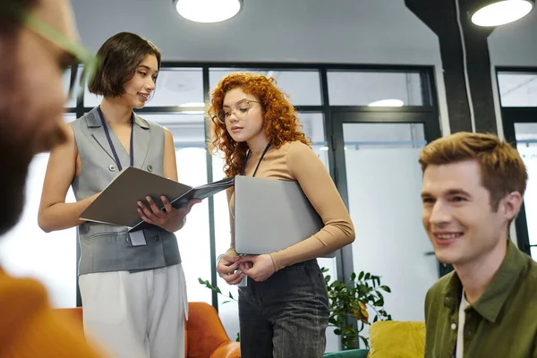 Mulheres de negócios jovens com documentos e laptop conversando perto de colegas borrados no espaço de escritório moderno — Fotografia de Stock