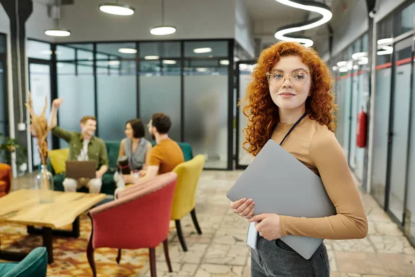 Redhead team lead with laptop near colleagues talking on blurred background, headshot — Stock Photo