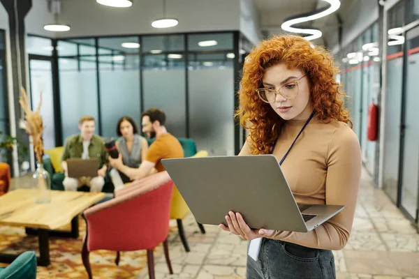 Pelirroja mujer de negocios mirando a la computadora portátil mientras el equipo hablando en un fondo borroso en la oficina moderna - foto de stock