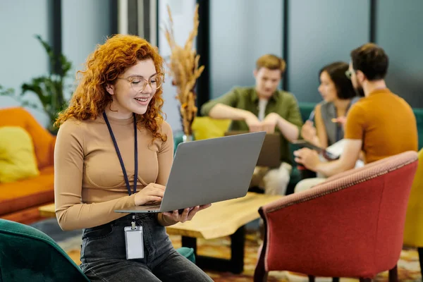 Gioiosa donna d'affari rossa con laptop vicino al team aziendale su sfondo sfocato in ufficio — Foto stock