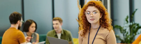 Redhead manager with laptop looking at camera near business team on blurred background in office — Stock Photo
