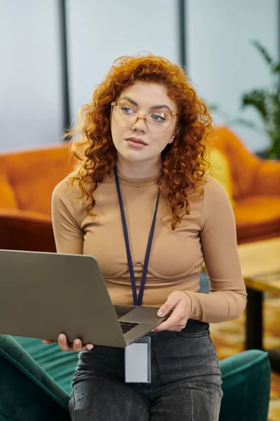 Pensive redhead businesswoman in eyeglasses looking away near laptop in contemporary office — Stock Photo