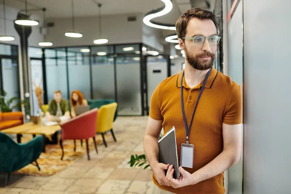 Entrepreneur barbu réfléchi avec des documents détournant les yeux, collègues flous travaillant dans le bureau — Photo de stock