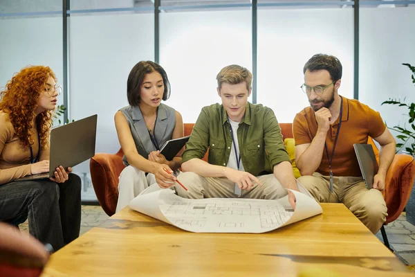 Молодой архитектор указывает на чертеж рядом с творческой командой бизнеса, работающей над запуском в офисе — стоковое фото