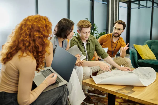 Redhead woman with laptop near colleagues talking near blueprint, teamwork in modern office — Stock Photo