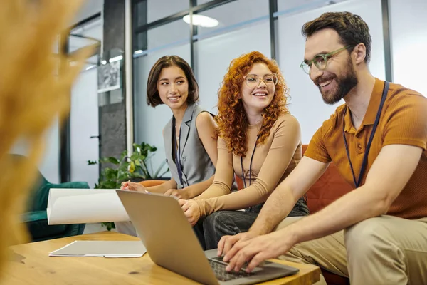 Positive bearded businessman working on laptop near smiling women with blueprint in coworking office — Stock Photo