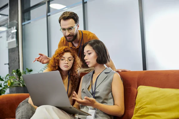 Capo squadra barbuto vicino alle donne d'affari che lavorano sul computer portatile in ufficio, pianificazione startup, lavoro di squadra — Foto stock