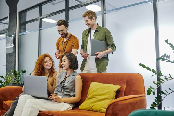 Gerentes jovens com laptop conversando no sofá perto de colegas de negócios no ambiente de co-trabalho moderno — Fotografia de Stock