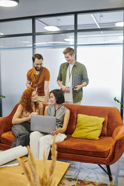 Hommes d'affaires positifs près de jeunes femmes avec ordinateur portable parlant sur le canapé dans le bureau moderne, projet de démarrage — Photo de stock