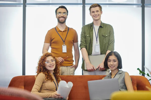 Профессиональный снимок бизнес-команды с ноутбуками и чертежом, улыбающейся в камеру в офисе — стоковое фото