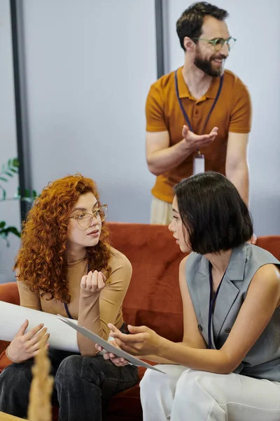 Geschäftsfrauen mit Dokumenten und Blaupause reden in der Nähe eines bärtigen Managers, der mit der Hand im Büro zeigt — Stockfoto