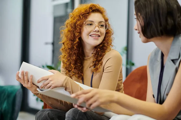 Redhead woman in eyeglasses holding blueprint and talking to business colleague in office — Stock Photo
