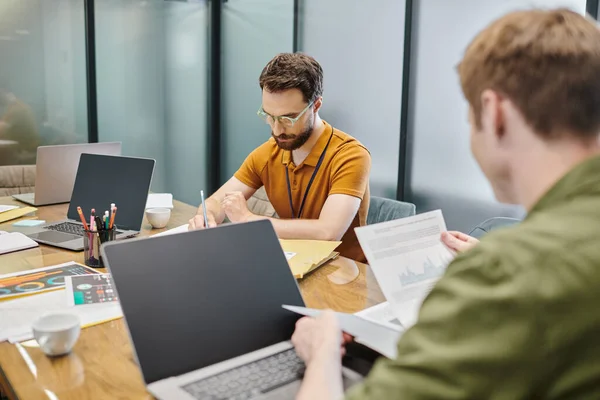 Bärtiger Geschäftsmann, der mit Dokumenten und Laptops nahe Kollegen schreibt, Arbeitsplatz in modernem Büro — Stockfoto