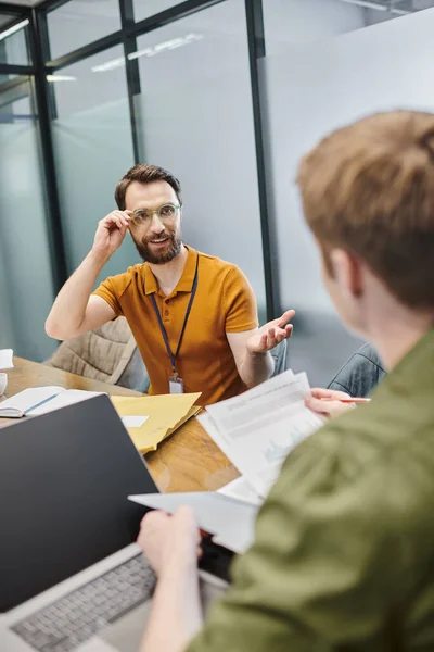 Bärtiger Geschäftsmann zeigt mit der Hand und spricht mit verschwommenem Kollegen mit Dokumenten in Laptopnähe — Stock Photo