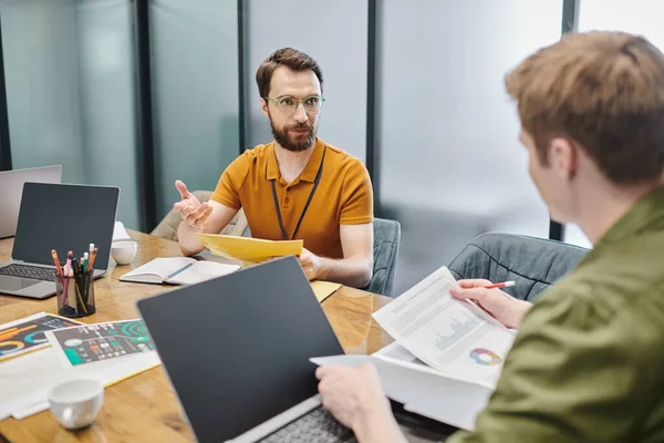 Bearded entrepreneur talking to colleague with documents near laptops and graphs at workplace — Stock Photo