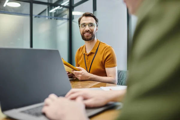 Bärtiger Geschäftsmann mit Umschlägen, lächelnd neben verschwommenem Kollegen, der auf Laptop im Büro tippt — Stockfoto