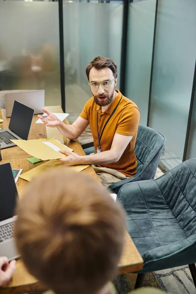 Bearded entrepreneur with envelopes talking to blurred business partners, laptops, modern workplace — Stock Photo