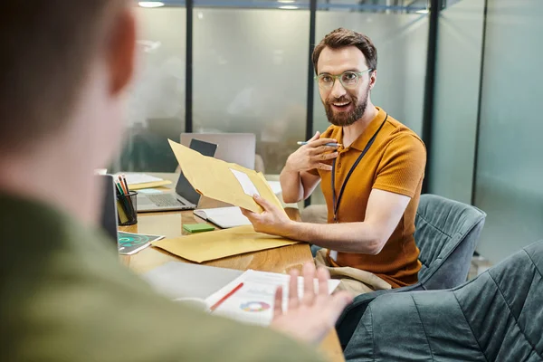 Bearded businessman with envelope talking to blurred colleague, laptops, documents, work environment — Stock Photo