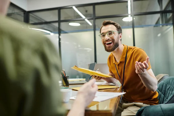 Cheerful bearded businessman with laptop talking to colleague on blurred foreground in modern office — Stock Photo