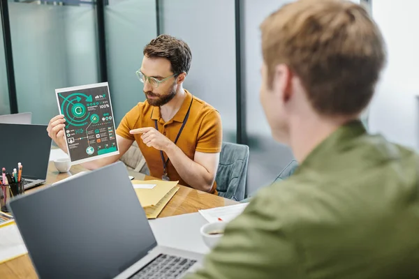 Bearded businessman pointing at infographics near laptops and blurred colleague in modern office — Stock Photo
