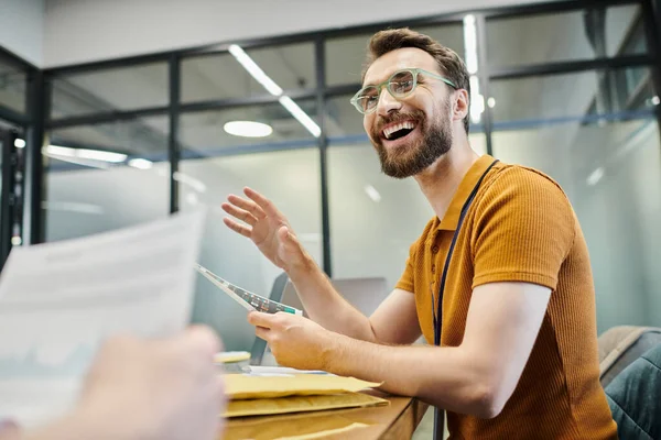 Excitado hombre de negocios barbudo con gráficos hablando con su colega en primer plano borroso en el lugar de trabajo - foto de stock
